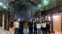 10 summer school students in blue hard hats stand in front of the MINERvA experiment.